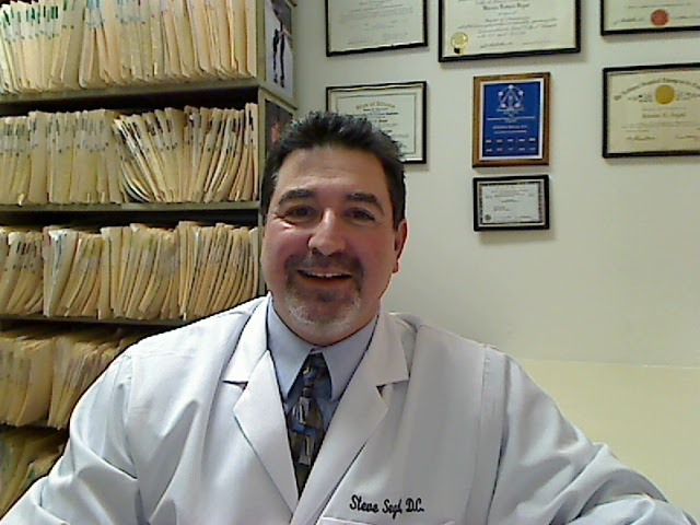 Dr. Steve Segal, Libertyville chiropractor, smiling while seated in his office wearing a white lab coat, with organized patient files and framed professional certificates on the wall behind him.